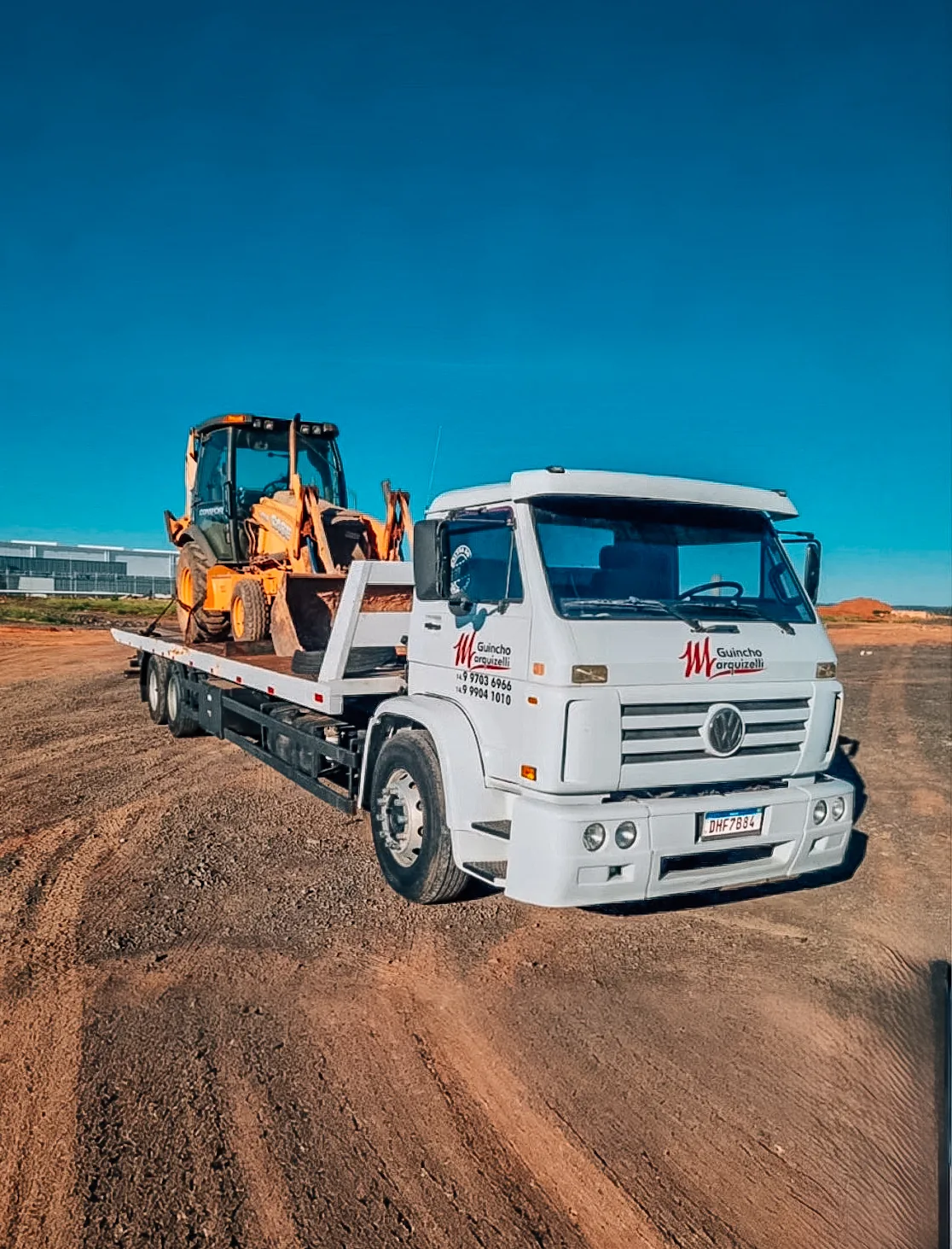 Caminhão plataforma branco transportando retroescavadeira